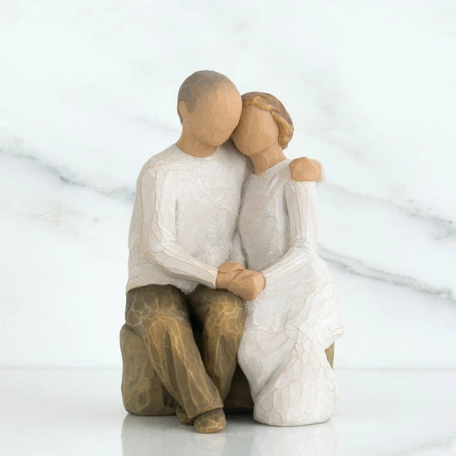 Willow Tree® Man and woman faceless figurines hugging one another sitting down on rocl - facing forward - woman is in white, man is in white and brown pants