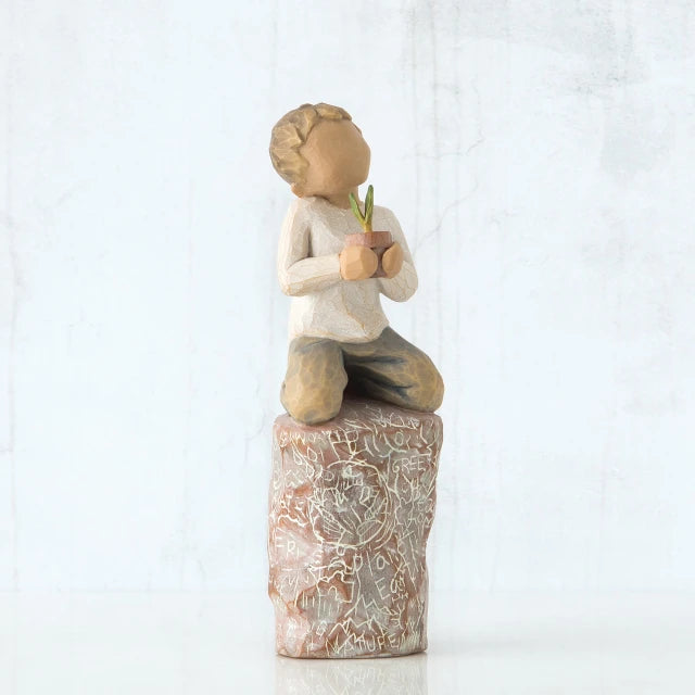 Willow Tree® Front view of boy with brown hair sitting on a stump while holding a small potted plant to his chest