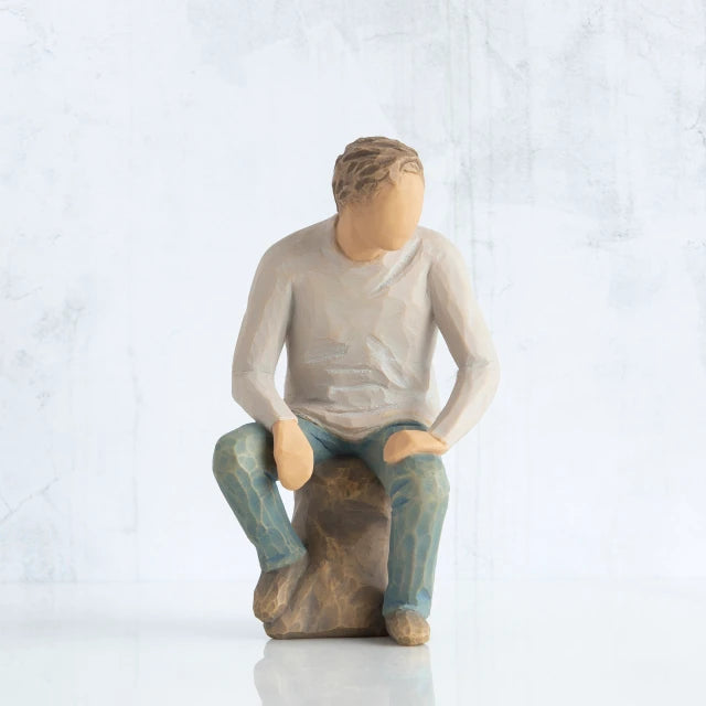 Willow Tree® Front view, adult male figure in light gray shirt and blue jeans, seated on gray rock