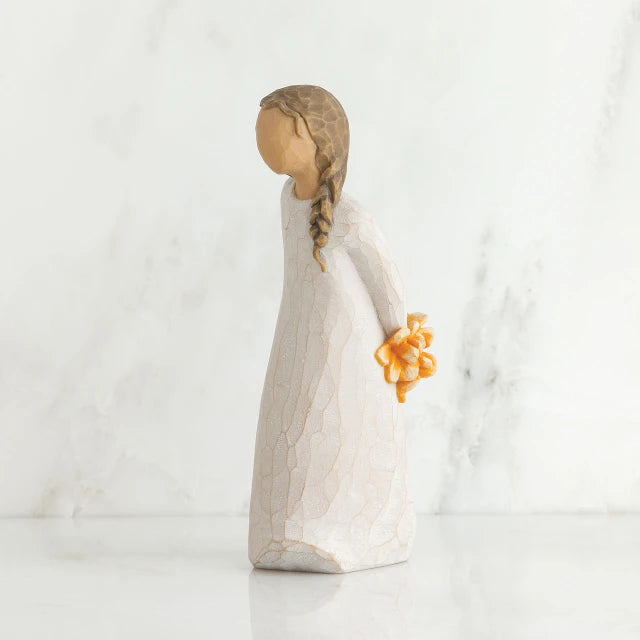 Willow Tree® Right side view of girl with brown hair in braid over right shoulder leaning forward as she holds orange flowers behind her back