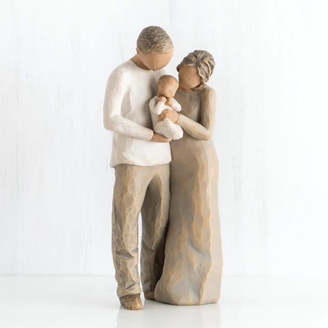 Willow Tree® Man and woman faceless figurines holding baby in white outfit - man is dressed in white shirt and brown pants, woman is dressed in brown dress