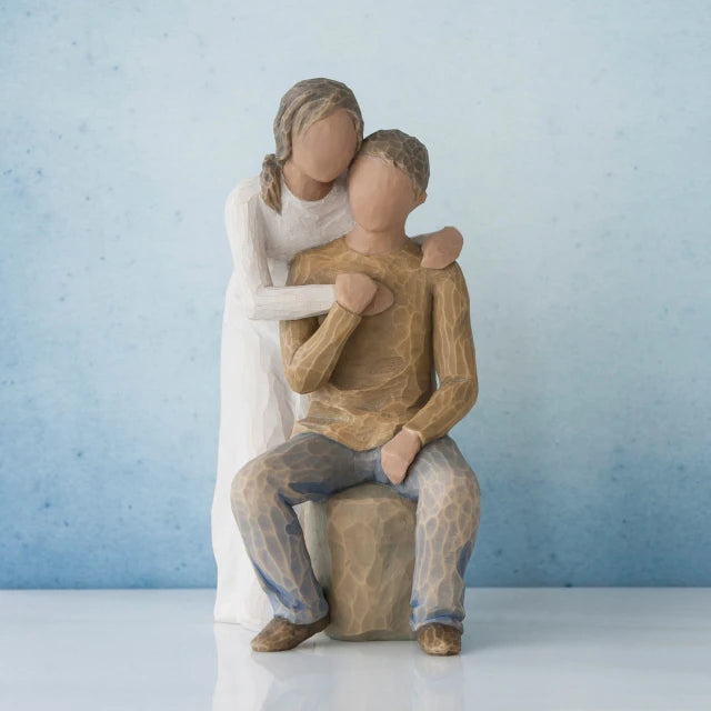 Willow Tree® Man figurine sitting on rock wearing brown shirt and blue jeans with woman figurine in white dress with arms wrapped around him - blue background